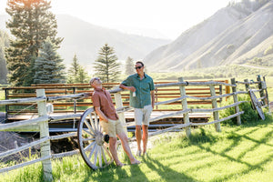 Two people standing near a wooden fence with mountains and trees in the background